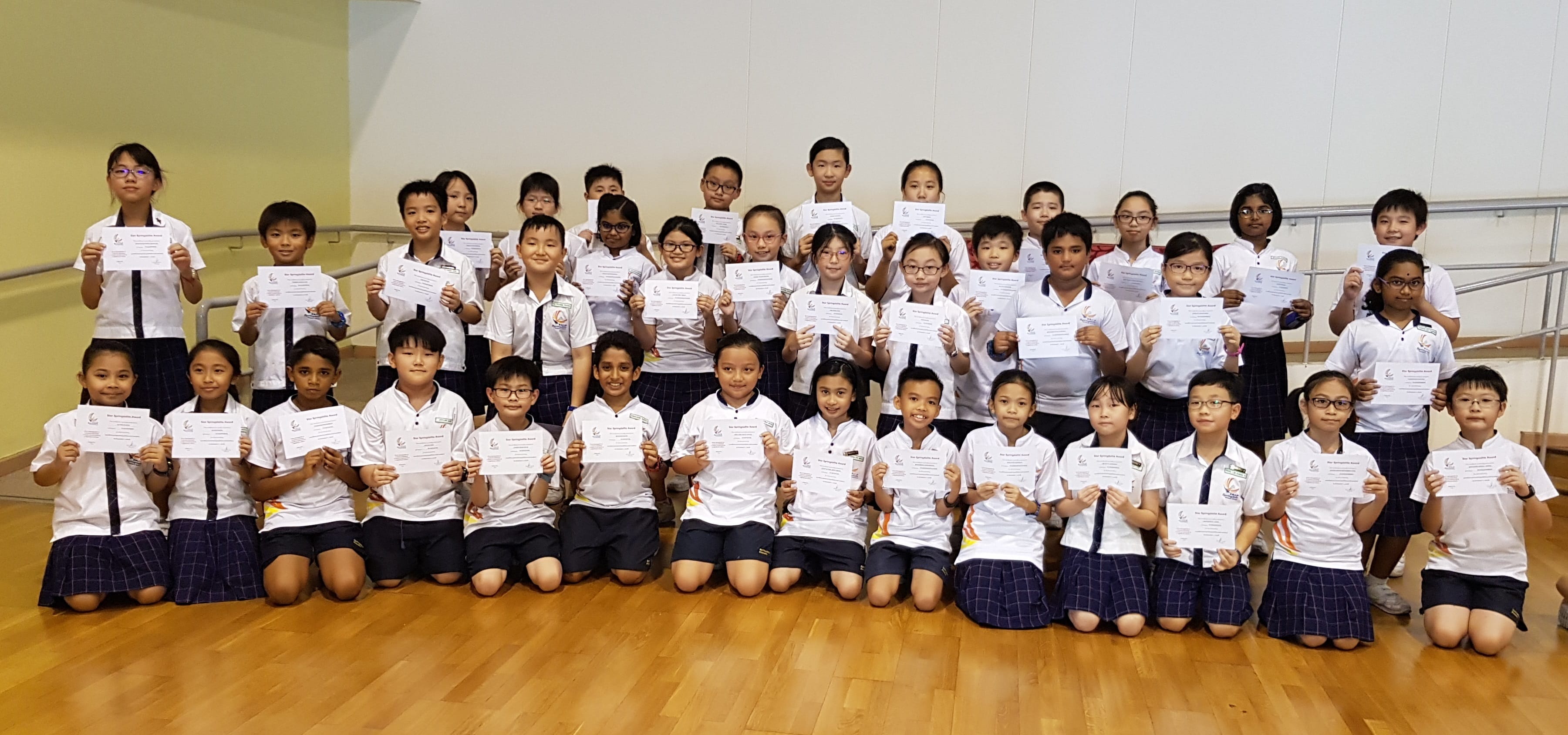 A large group of school children in white uniforms pose in three rows holding certificates indoors.